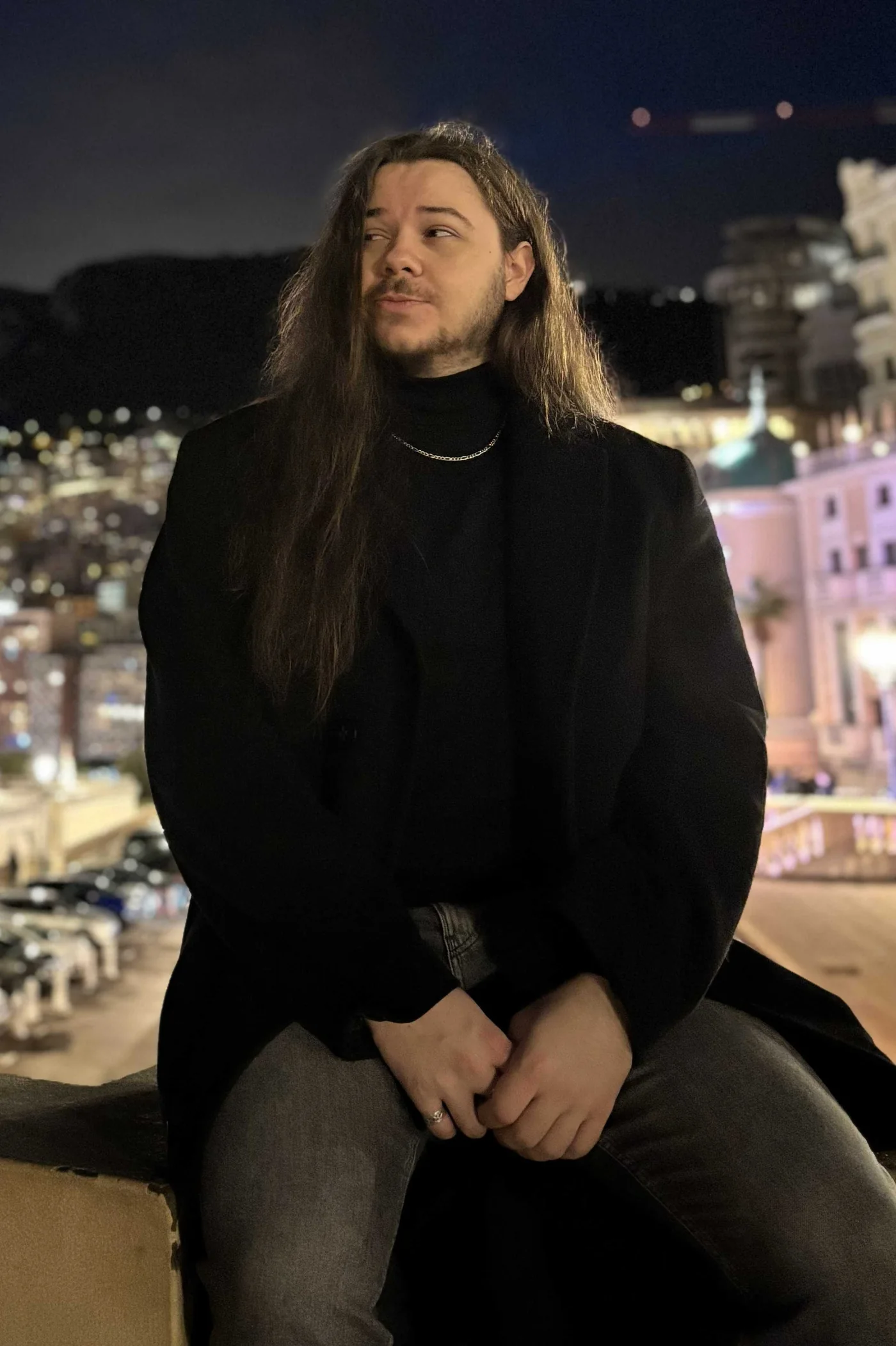 Martin Skořupa in a black coat, sitting on a low wall with illuminated buildings behind him